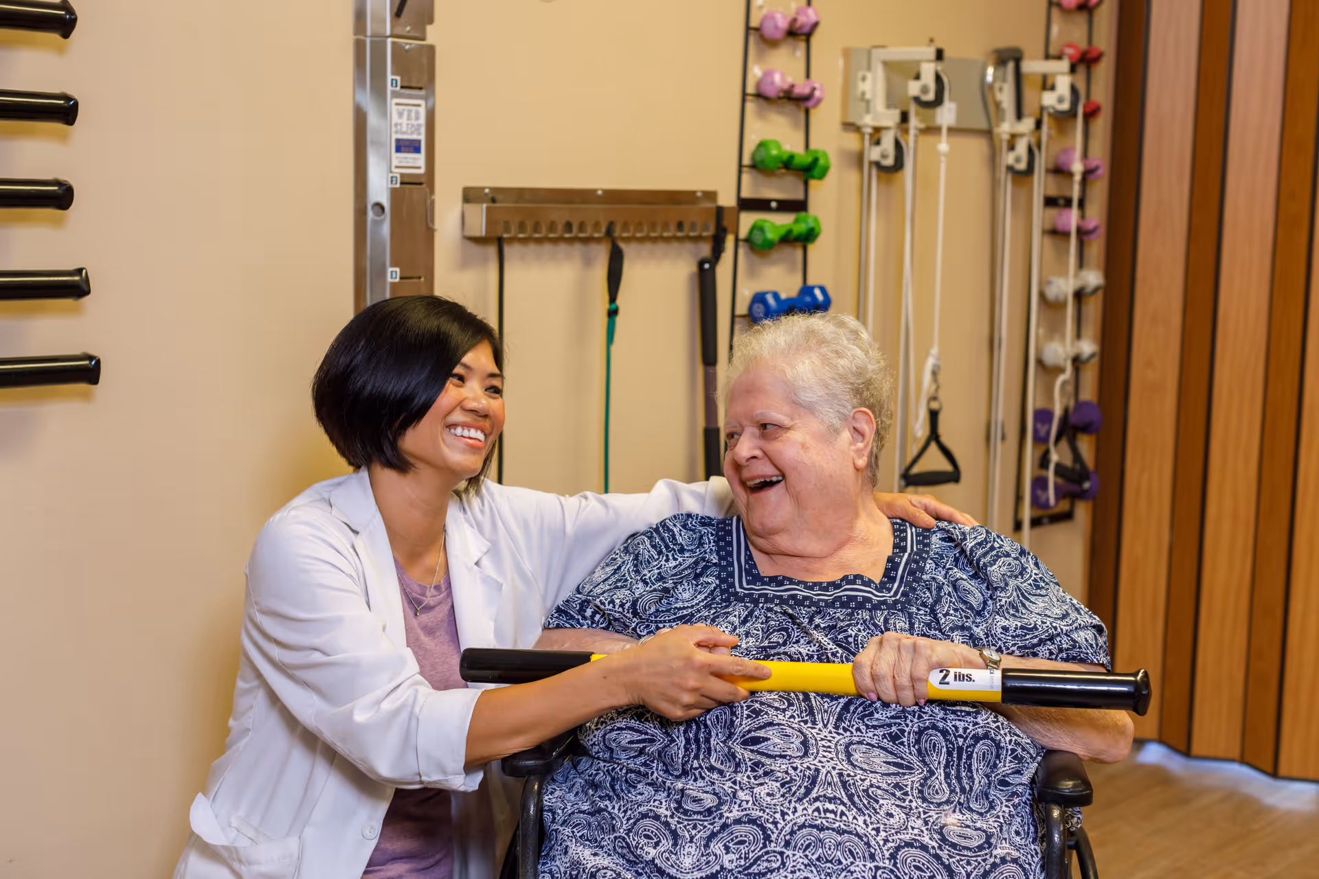 A healthcare professional in a white coat smiles and supports an elderly woman in a wheelchair as they hold a yellow exercise bar labeled '2 lbs.' They are in a room with various exercise equipment on the walls, including dumbbells and resistance bands.