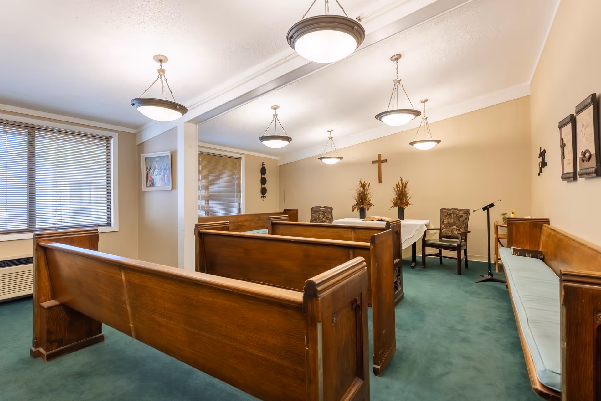 Interior of a small chapel or prayer room with wooden pews, a green carpet, a table covered with a white cloth, two floral arrangements, a wooden cross on the wall, and framed religious artwork. The room is lit by multiple ceiling lights and has windows with blinds.