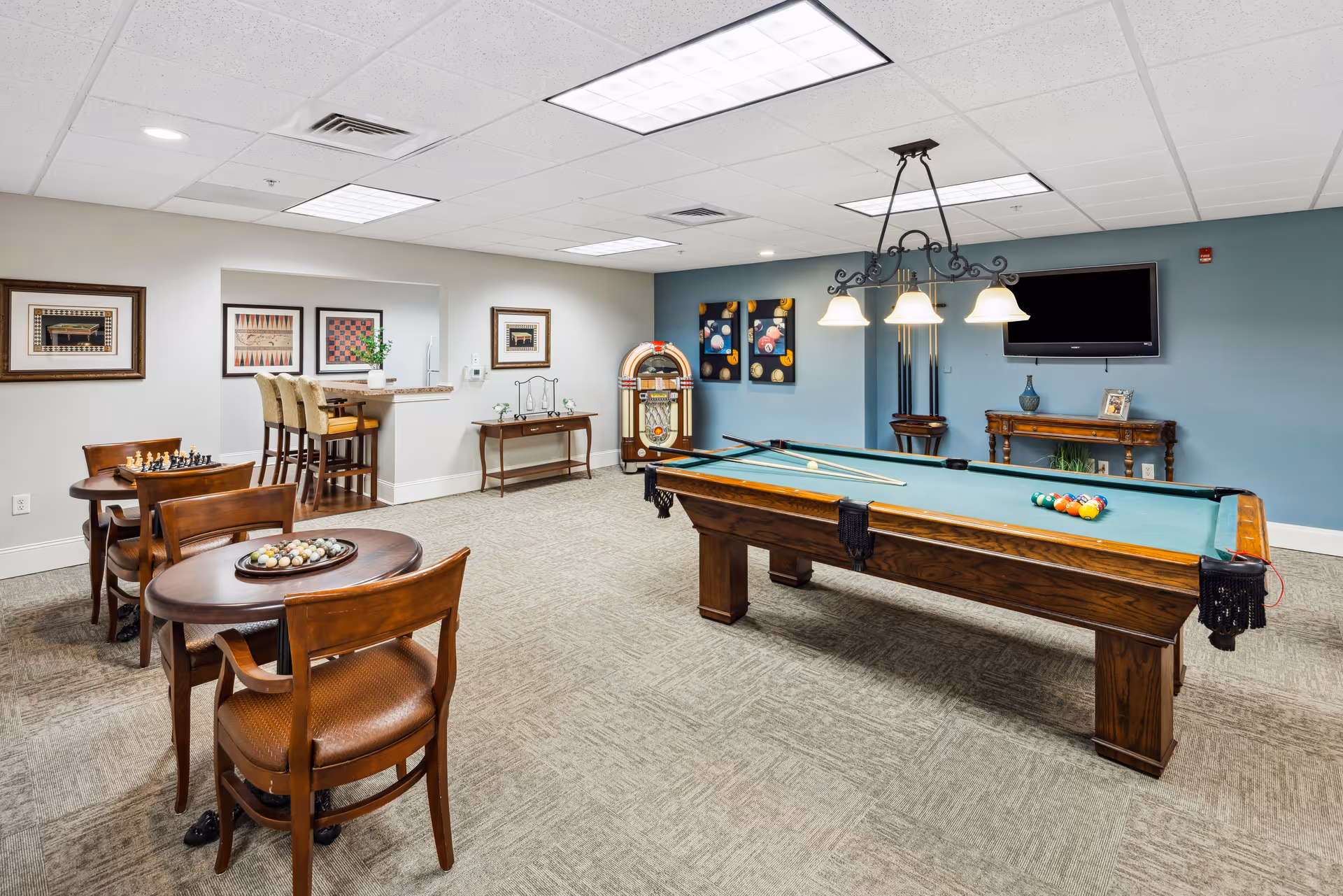 A recreational room with a pool table in the center, a hanging light fixture above it, and a wall-mounted TV on a blue accent wall. There is a vintage jukebox in the corner, a small table with pool cues, and two small round tables with chairs, one with a chess set and the other with a decorative bowl. The room has framed artwork on the walls and a carpeted floor.