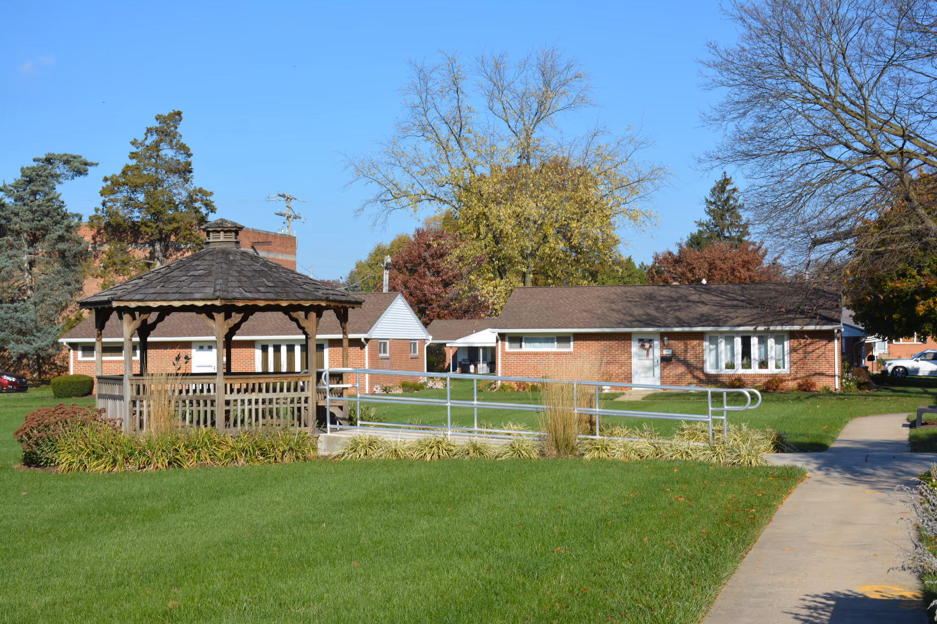 Outdoor view of a senior living facility with a wooden gazebo surrounded by greenery and a paved walkway. In the background, there are single-story brick buildings with windows and doors, trees with autumn foliage, and a clear blue sky.
