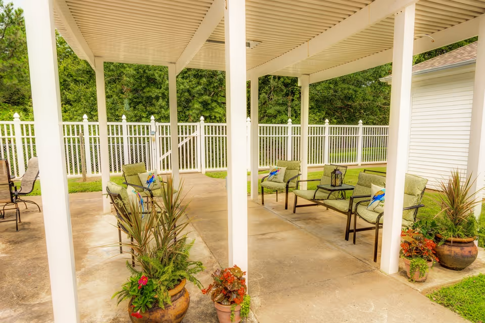 Outdoor covered patio area with green cushioned chairs and small tables, surrounded by potted plants and a white fence with green trees in the background.