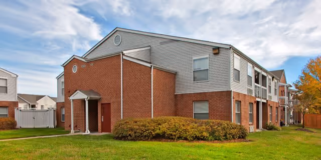 Exterior view of a two-story residential building with brick and siding walls, surrounded by green grass and bushes under a partly cloudy sky.