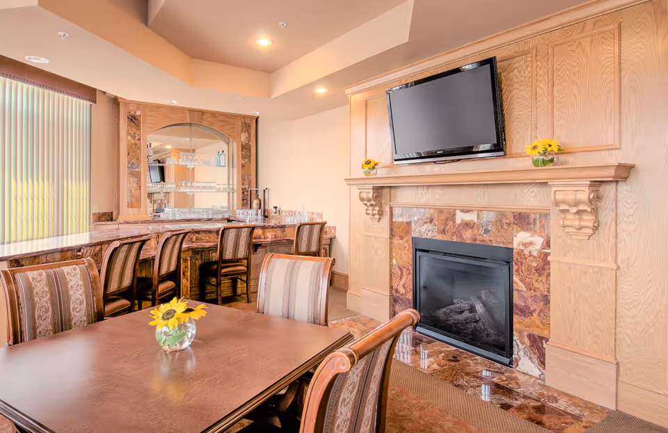 A cozy interior room featuring a wooden dining table with a small vase of sunflowers, upholstered chairs, a bar area with high chairs and glassware, a fireplace with a marble surround, and a flat-screen TV mounted above the mantel.