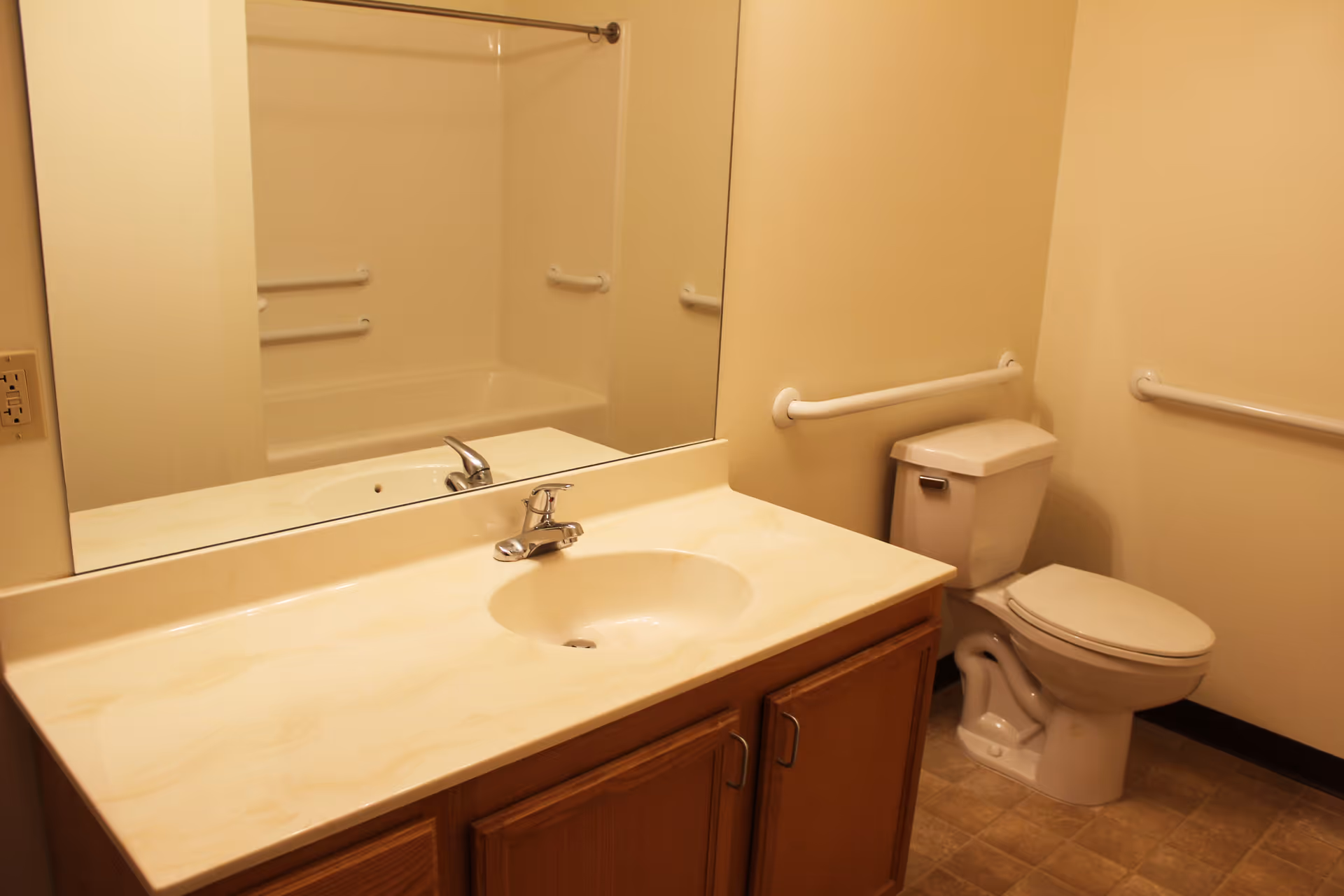A bathroom with a beige countertop sink, wooden cabinets below, a large mirror above the sink, a toilet with grab bars on the walls, and a bathtub with a shower curtain rod and grab bars visible in the reflection.