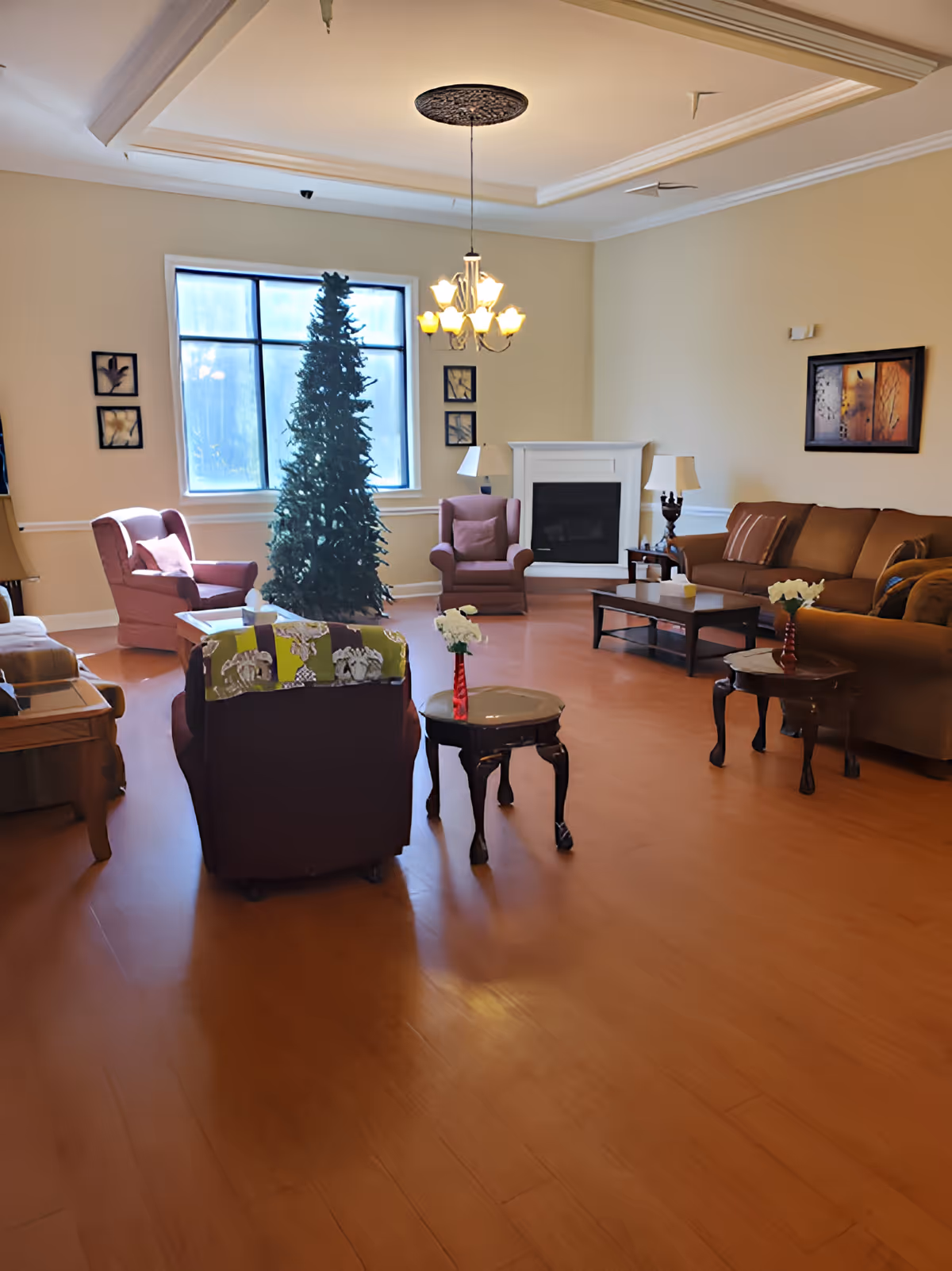 A spacious living room with wooden flooring, featuring a brown sofa, several armchairs, two small tables with red vases holding white flowers, a fireplace, a large window, and a tall, undecorated Christmas tree in the center. The walls are light-colored with framed artwork, and a chandelier hangs from the ceiling.