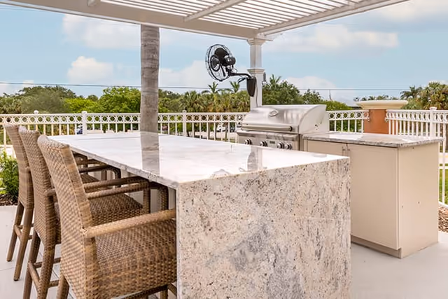 Outdoor patio area with a granite countertop bar and four wicker bar stools under a pergola. A stainless steel grill and a mounted fan are visible in the background, along with a white railing and greenery.