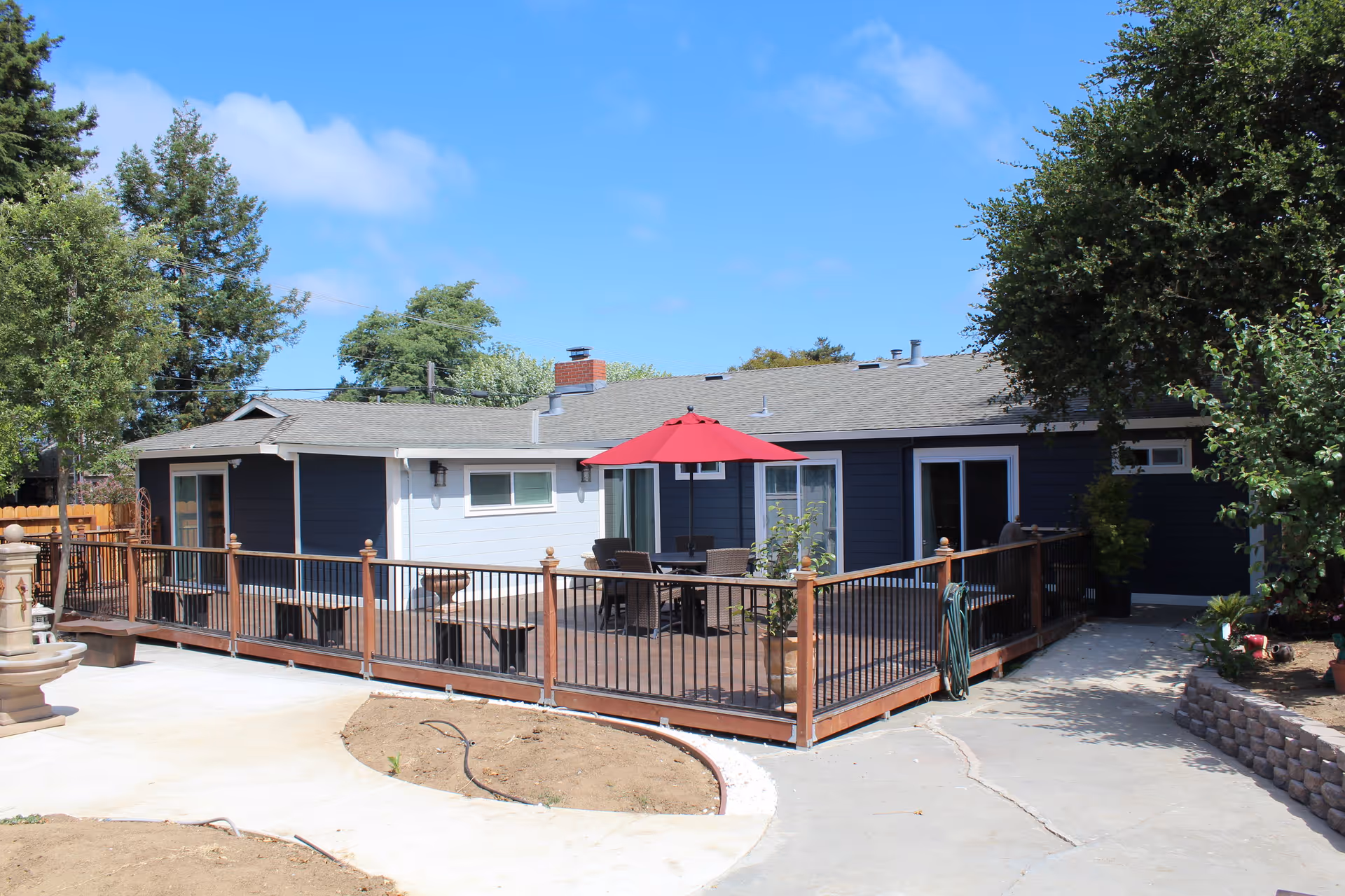 Outdoor patio area of a single-story building with dark blue and white exterior walls. The patio is fenced with wooden and metal railings and features a table with chairs under a red umbrella. Surrounding the patio are trees, plants, and a concrete pathway.