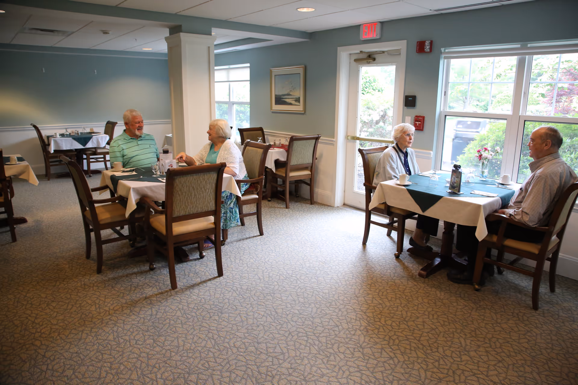 Two elderly couples sitting at separate tables in a well-lit dining room with large windows, having a conversation. The room has carpeted floors, light blue walls, and tables set with tablecloths, cups, and utensils.