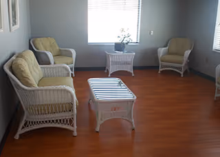 A small sitting area with white wicker furniture including a loveseat, two armchairs, and a coffee table on a wooden floor. There is a window with blinds and a small plant on a side table between the chairs.