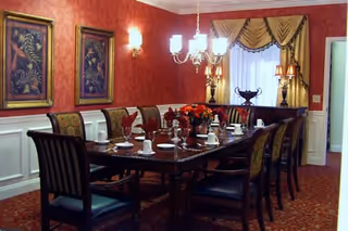 Formal dining room with a long polished table set with dishes and cups, upholstered chairs, chandelier, framed artwork and red patterned walls.