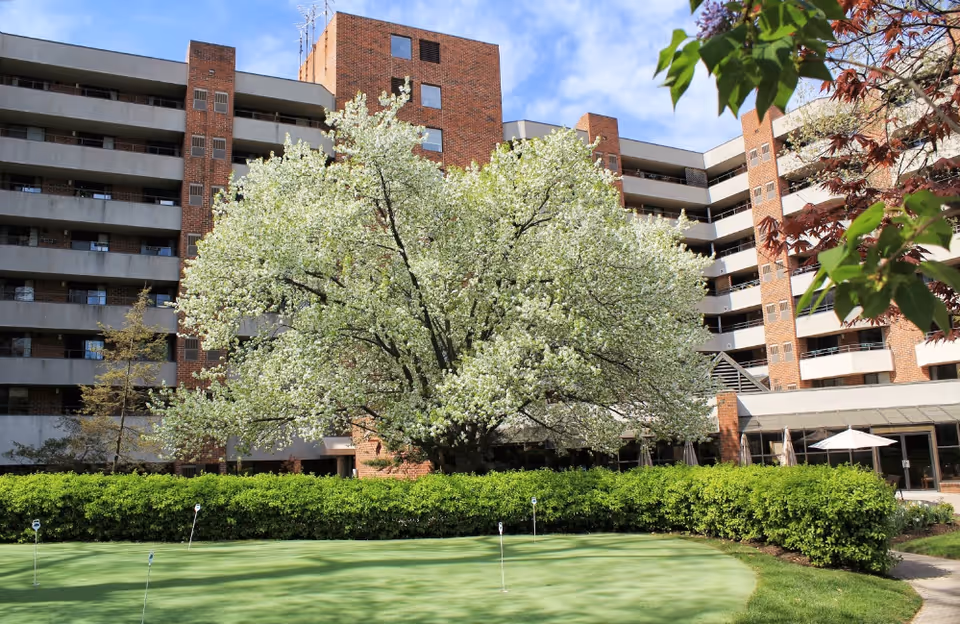 Multi-story brick building with balconies, a large flowering tree, and a small putting green in the foreground.