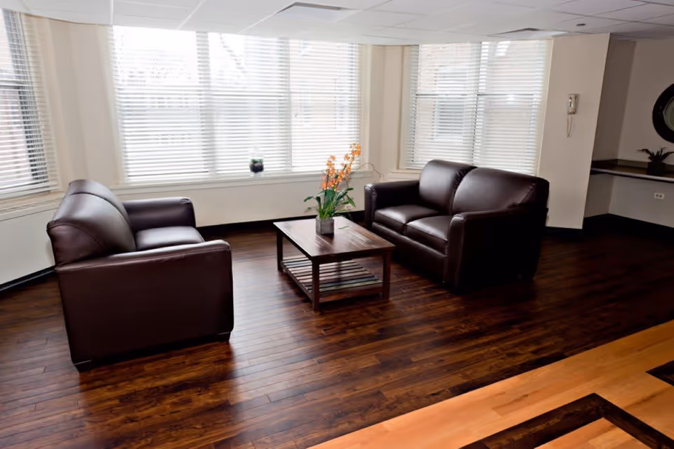 A bright living room area with two dark brown leather sofas facing each other and a wooden coffee table with a potted plant on it, set against large windows with white blinds and dark wooden flooring.