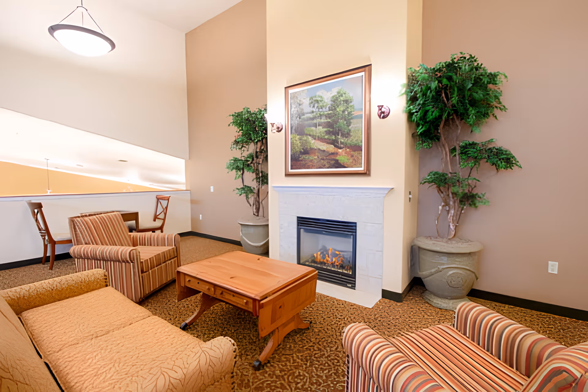 A cozy senior living room with a beige couch and two striped armchairs arranged around a wooden coffee table. A fireplace with a lit fire is centered on the wall, above which hangs a framed painting of a landscape with trees. Two large potted plants flank the fireplace. In the background, there is a small table with two wooden chairs near a half wall. The room has warm beige and brown tones with a patterned carpet and a ceiling light fixture.