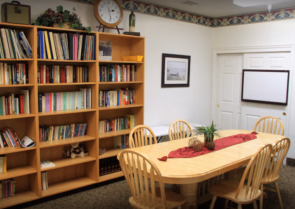 A cozy room with a wooden table surrounded by six wooden chairs. The table has a red runner with decorative items and a small plant on it. Behind the table, there is a large wooden bookshelf filled with books and some decorative items. A clock and a green bottle are placed on top of the bookshelf. On the wall, there is a framed picture and a whiteboard mounted on white double doors. The room has a patterned carpet and wallpaper border near the ceiling.