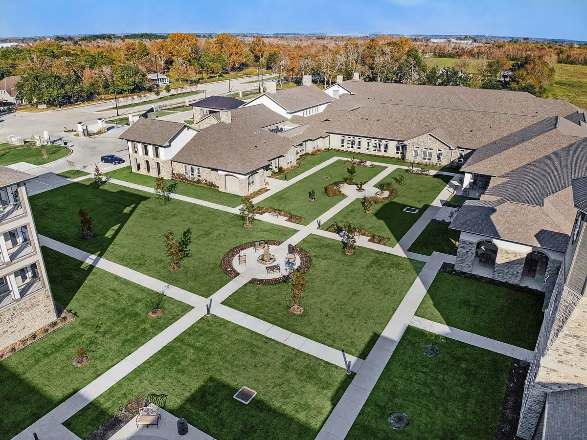 Aerial view of The Lodge at Pine Creek showing a large courtyard with green lawns, paved walkways, benches, and a circular seating area with chairs and a small fountain. Surrounding the courtyard are multiple connected buildings with stone and brick exteriors under a clear blue sky.