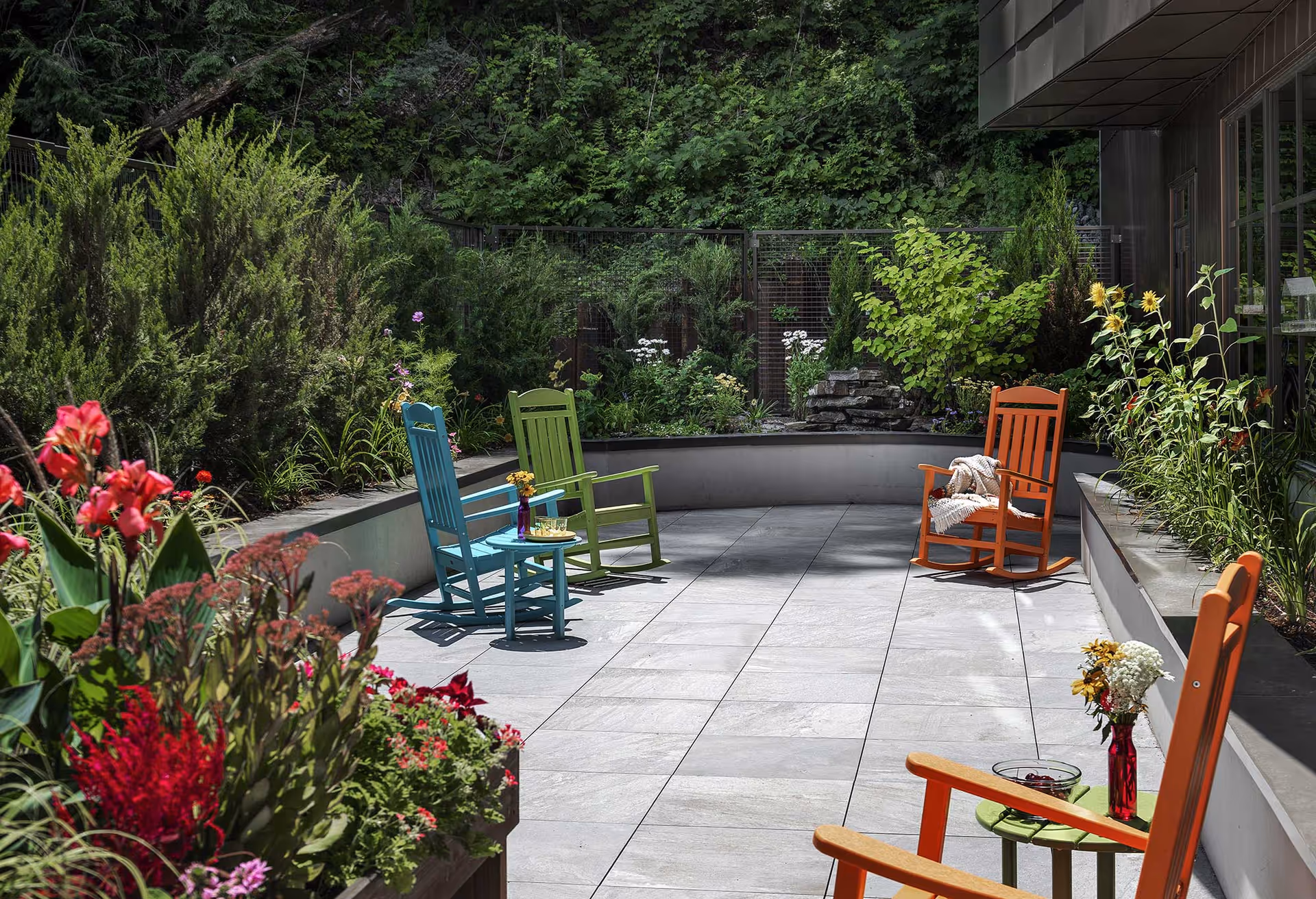 Sunlit outdoor courtyard patio with colorful rocking chairs, small tables and surrounding plants and greenery.
