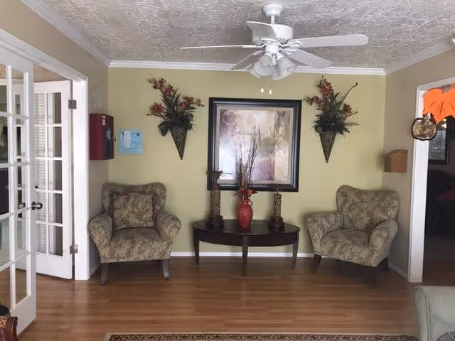 A cozy sitting area in a senior living facility with two patterned armchairs on either side of a dark wooden console table. The table holds a red vase with decorative branches and two candle holders. Above the table hangs a large framed abstract painting, flanked by two wall-mounted floral arrangements. The room has light green walls, a white ceiling fan, and wooden flooring. To the left, there are glass-paneled double doors, and to the right, an open doorway leads to another room.