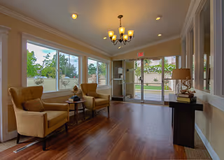 A well-lit interior common area with large windows showing an outdoor view. The room features two beige armchairs with a small round table between them, a chandelier hanging from the ceiling, and a console table with a lamp and decorative items near the entrance door.