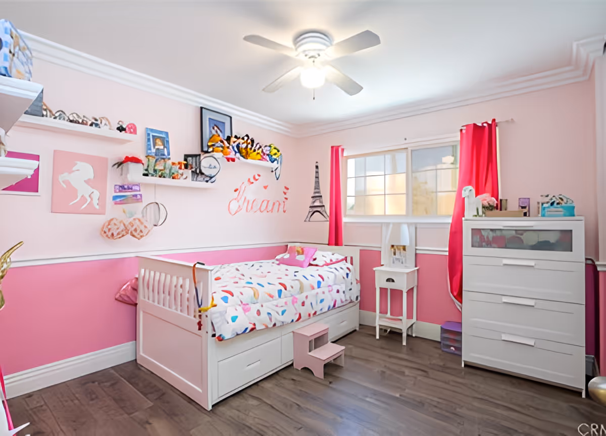 A bright and colorful bedroom with pink walls and white trim. The room features a white daybed with a colorful patterned comforter and a small pink step stool in front of it. Above the bed, there are white floating shelves holding various stuffed animals, framed pictures, and decorative items. The name 'Ivani' is written on the wall in red cursive letters next to a drawing of the Eiffel Tower. A white dresser and a small white side table are positioned near a window with pink curtains. The room has a ceiling fan with a light and wooden flooring.