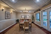 A dining room with a rectangular wooden table set for six people. The room has beige walls with white trim, carpeted floor, two framed pictures on the far wall, a chandelier hanging from the ceiling, wall sconces providing additional lighting, and large windows and glass doors on the right side leading to an outdoor area.