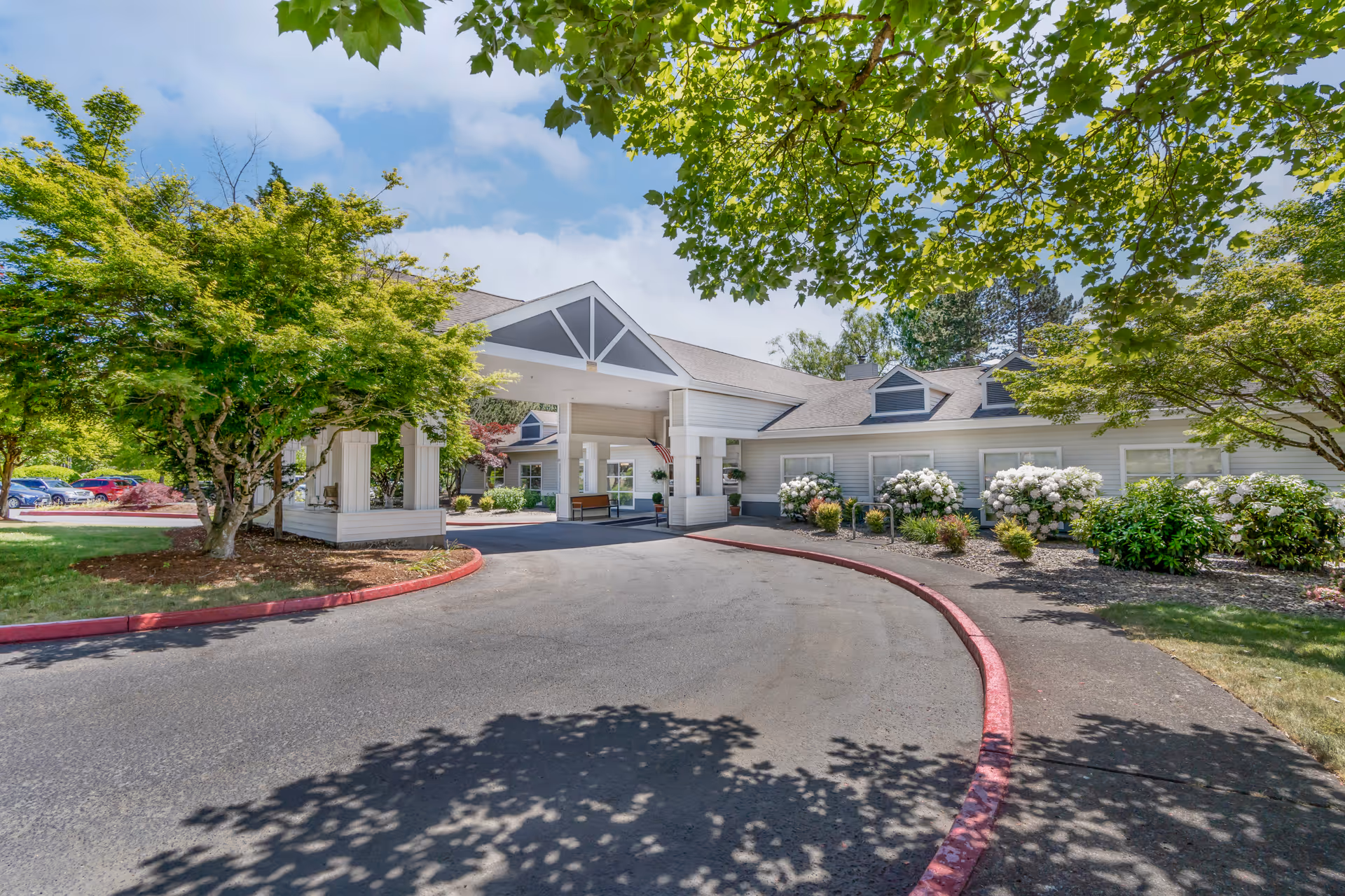 Exterior view of Brookdale Mt. Hood facility entrance with a covered drop-off area, surrounded by green trees, bushes with white flowers, and a clear blue sky.