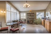A bright dining area with large windows covered by white blinds, featuring two round glass-top tables each surrounded by four chairs with red cushions. A chandelier hangs from the ceiling, and there is a wooden cabinet with shelves and a potted plant near the back wall. The room has light-colored walls and flooring.