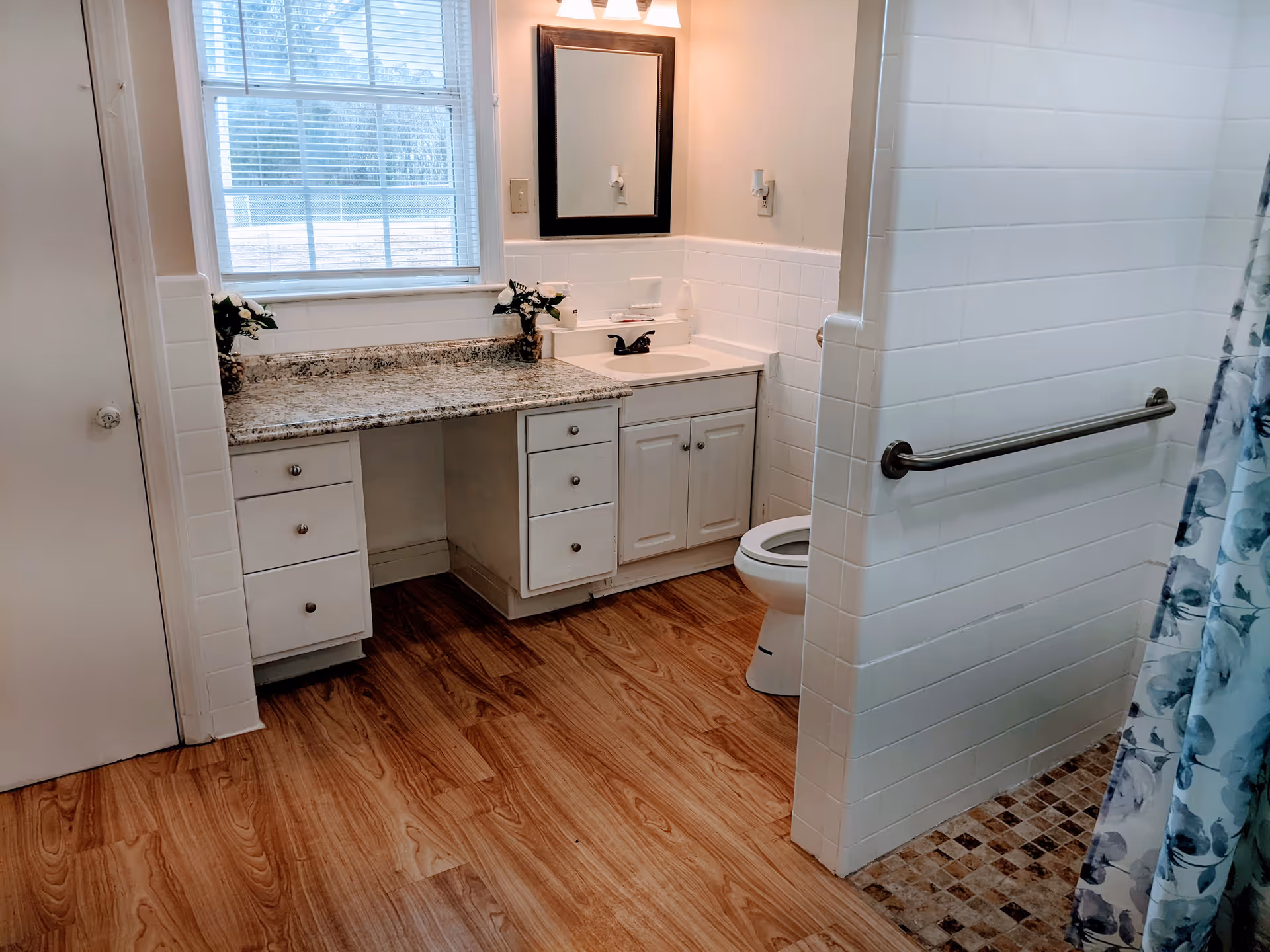 Accessible bathroom with a vanity and sink, toilet, walk-in shower with grab bar and floral curtain, and wood-look flooring.
