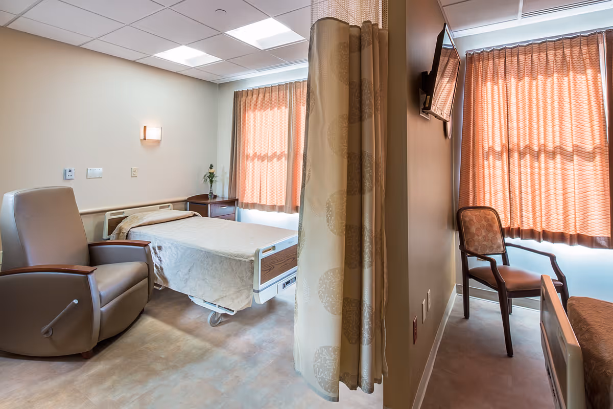 A private senior care room with a hospital bed, reclining chair, side table, wall-mounted TV and privacy curtain.