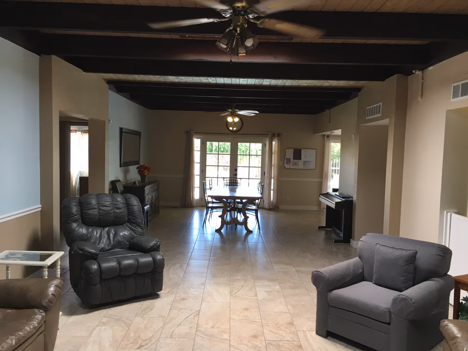 A spacious living area with tiled floors and wooden ceiling beams. The room features a black leather recliner, a gray armchair, a wooden dining table with chairs near large windows with curtains, and a piano against the right wall. Ceiling fans with lights are mounted on the ceiling.