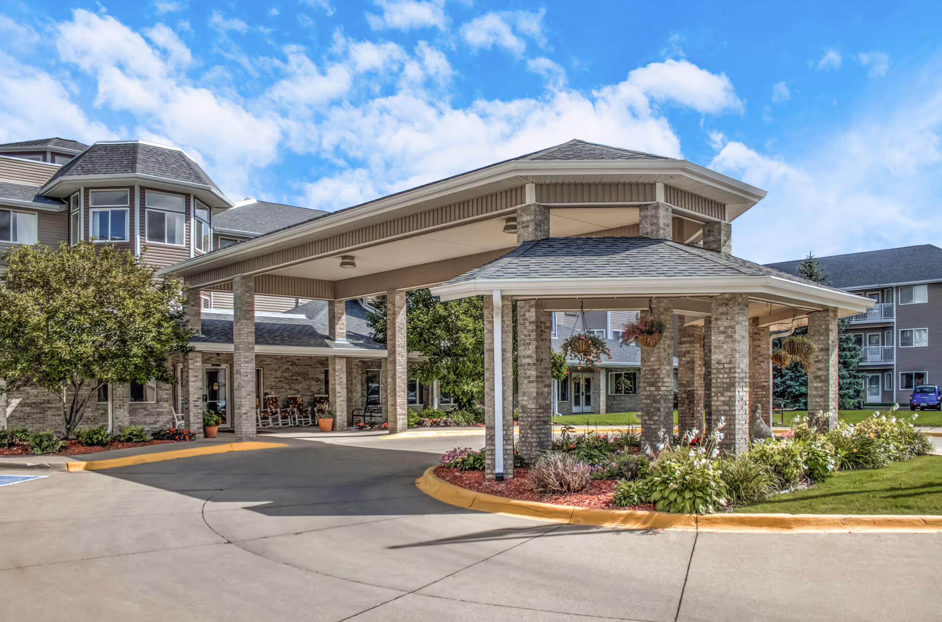 Covered porte-cochere and main entrance of a senior living facility with brick columns, hanging plants, landscaped beds and a driveway.