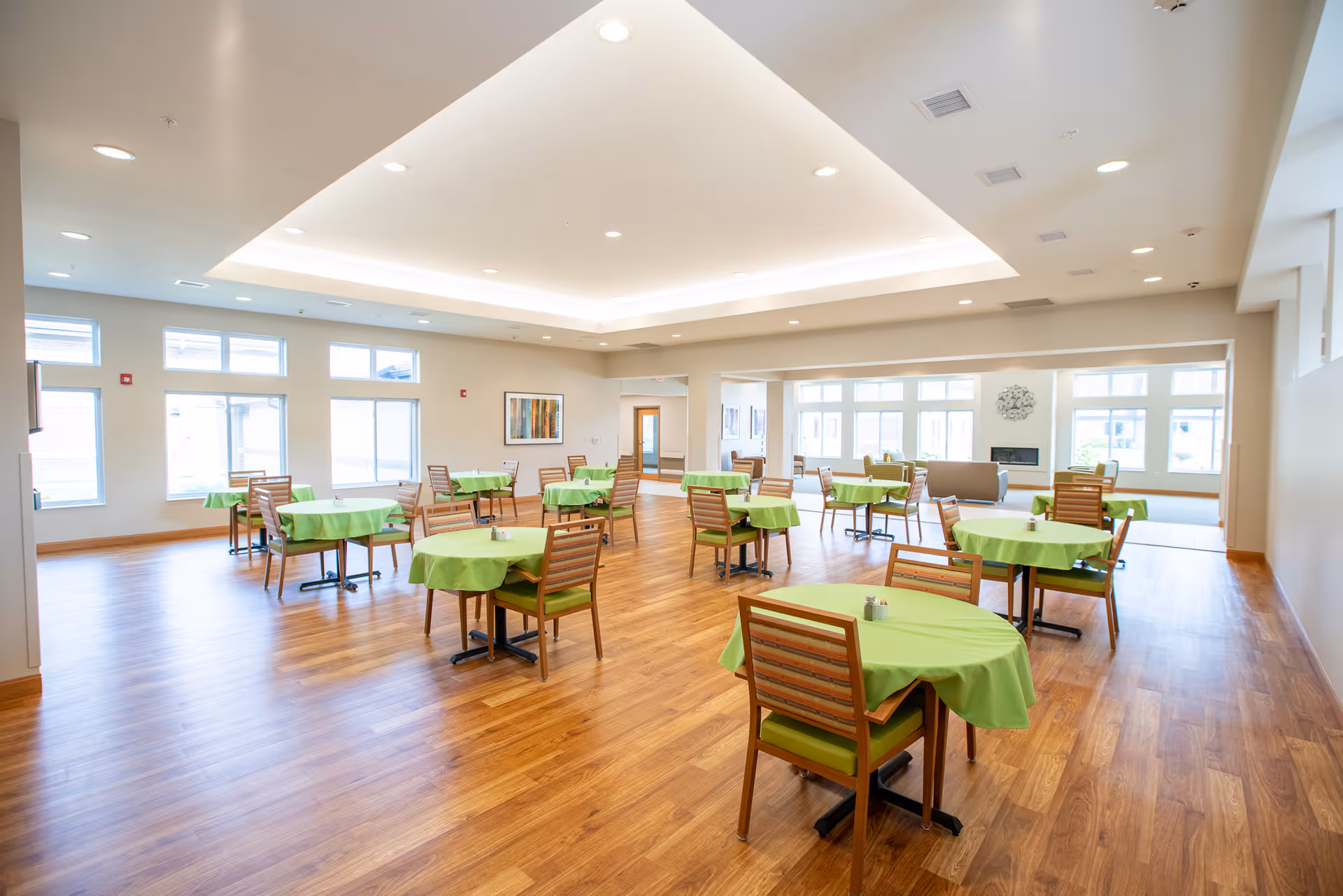 A spacious, well-lit dining room with multiple round tables covered in green tablecloths, each surrounded by wooden chairs with green cushions. The room has large windows allowing natural light to fill the space, light-colored walls, and a wooden floor. There is a seating area with a couch and a fireplace visible in the background.