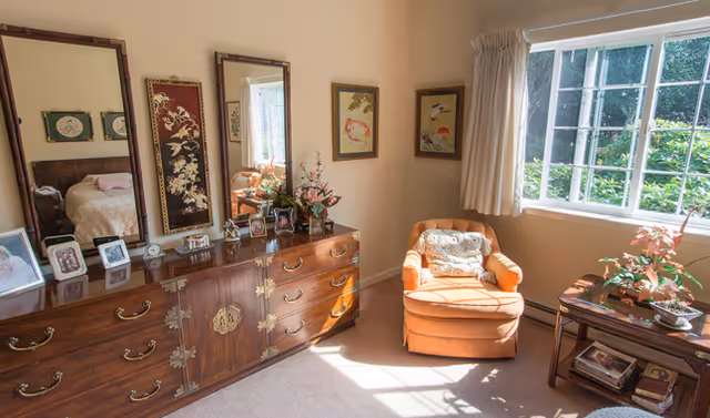 A cozy room with a large wooden dresser featuring ornate handles and three mirrors above it. The dresser top holds several framed photographs and decorative items. To the right, an orange upholstered armchair with a patterned cushion sits next to a window with white curtains, letting in natural light. A small wooden side table with a plant and other decorative items is beside the chair. The walls are adorned with framed artwork.