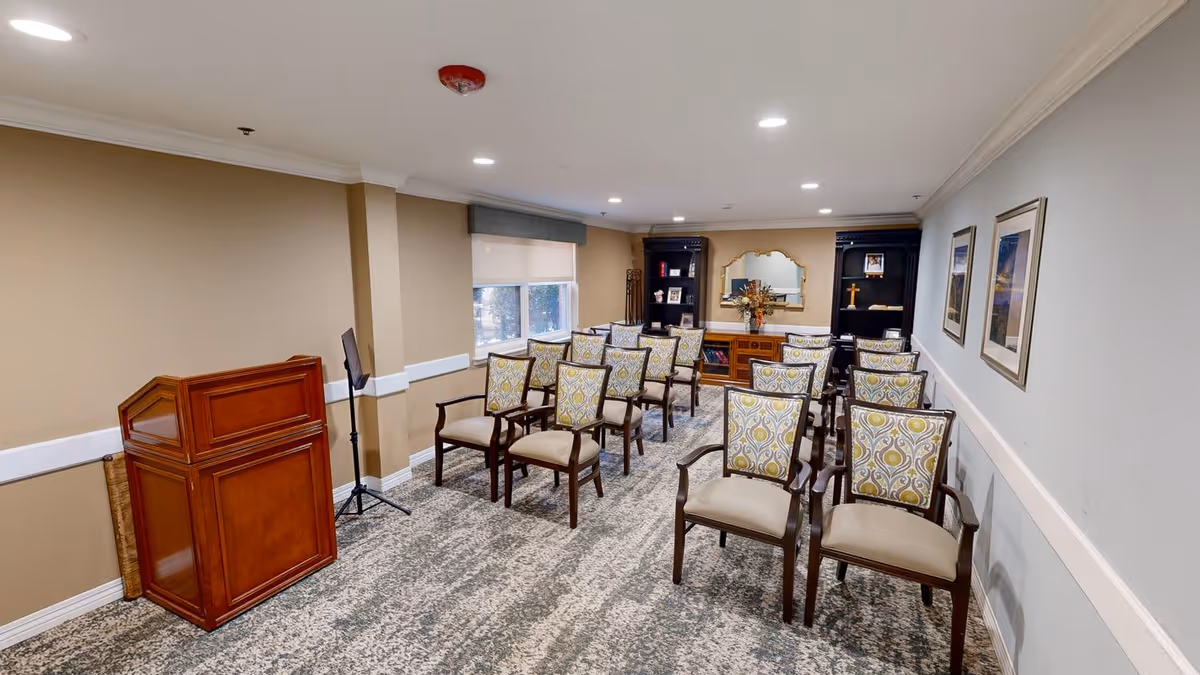 A small meeting or presentation room with rows of wooden chairs featuring patterned cushions arranged facing a wooden podium. The room has beige walls, carpeted floor, recessed ceiling lights, a window with a blind, and framed pictures on the wall. At the far end, there are two black bookshelves with decorative items and a large mirror in the center.