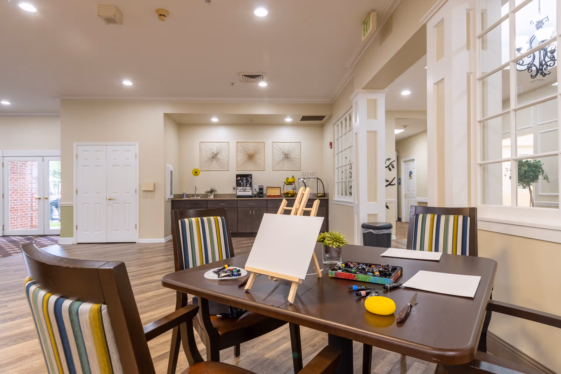 A well-lit interior room with a wooden table set up for painting activities, including blank canvases on easels, paint tubes, brushes, and paper. The room has striped cushioned chairs around the table, wood flooring, and a counter with a coffee machine and decorative wall art in the background. Large windows and doors allow natural light into the space.