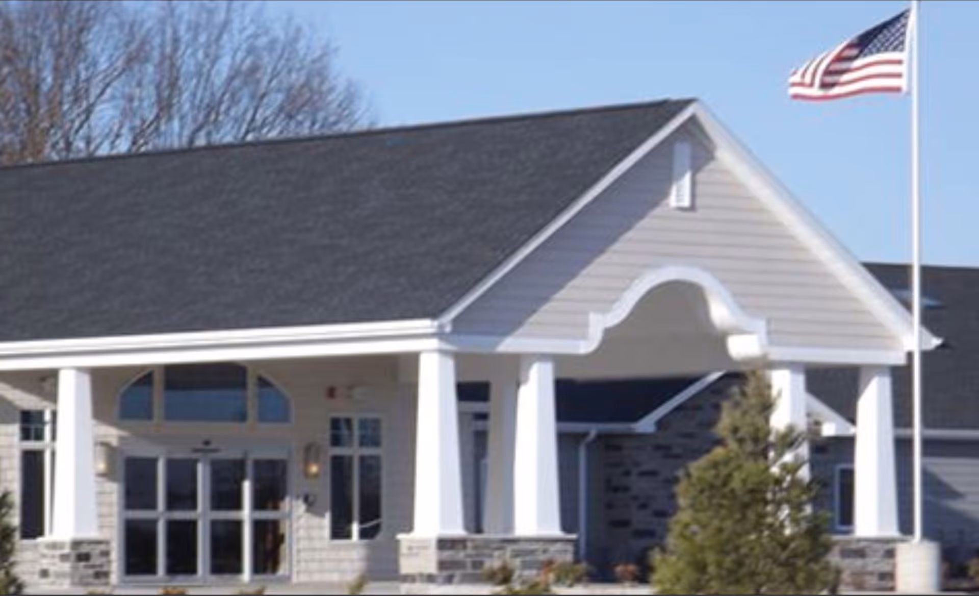 Exterior front entrance of a senior living facility with a covered porch supported by white columns, stone accents at the base, and an American flag flying on a flagpole to the right.