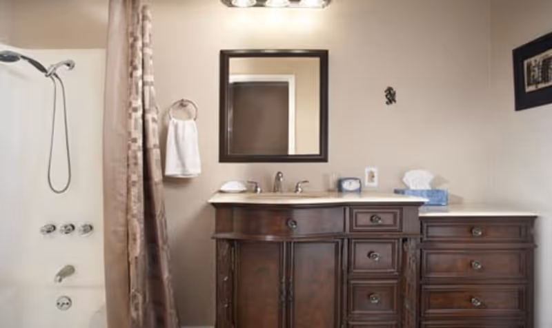 A bathroom with a dark wooden vanity featuring multiple drawers and a white countertop. Above the vanity is a rectangular mirror with a dark frame and a light fixture with three bulbs. To the left is a bathtub with a showerhead and a beige shower curtain. A white towel hangs on a ring next to the mirror, and a tissue box and clock are on the countertop.