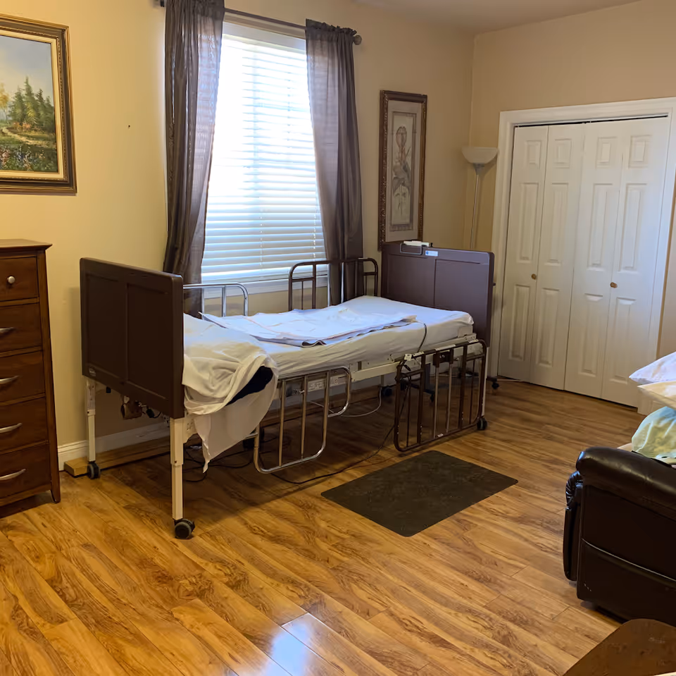 A senior care bedroom with a hospital-style bed positioned near a window with brown curtains. The room has wooden flooring, a wooden dresser, a floor lamp, framed artwork on the walls, and a closet with white double doors.