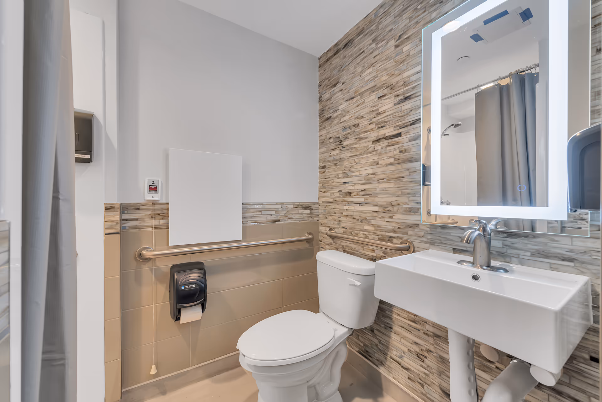 Accessible bathroom with a toilet, wall-mounted sink and illuminated mirror, grab bars and a shower curtain.