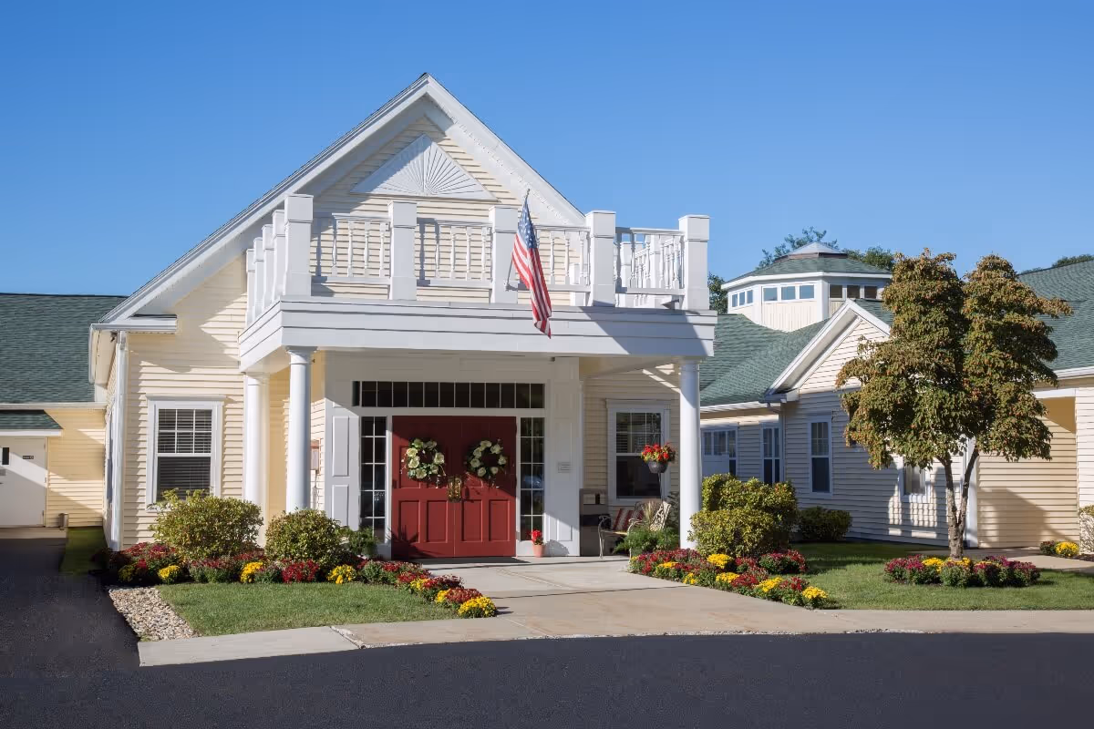 Front exterior view of a senior living facility named The Atrium at Veronica Drive, featuring a light yellow building with a green roof, a covered entrance with white columns, red double doors decorated with wreaths, an American flag, and well-maintained landscaping with colorful flowers and bushes.