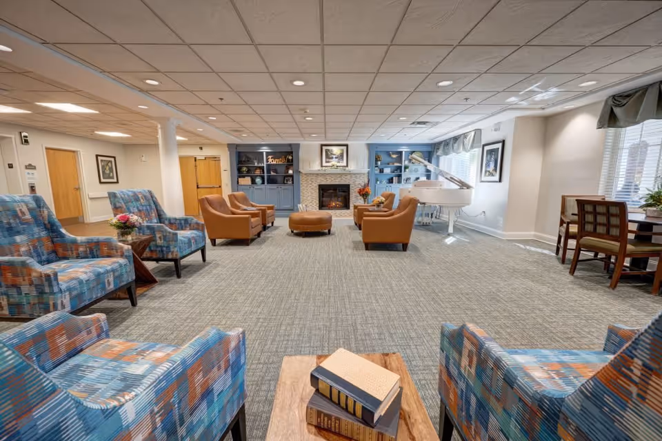 A spacious and well-lit common area with patterned blue and orange armchairs, tan leather chairs, a round ottoman, a white grand piano near large windows, built-in blue shelves flanking a fireplace, and a small table with chairs in the corner.