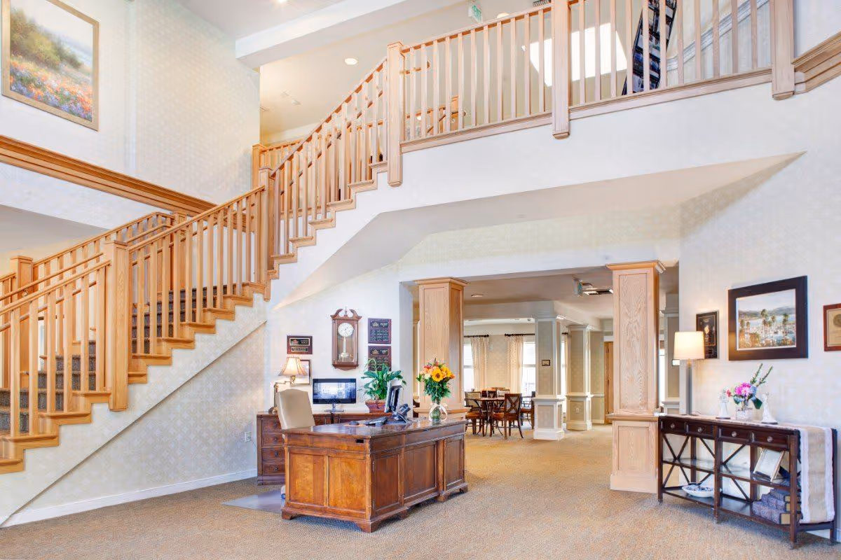 Interior view of a senior living facility lobby with a wooden reception desk, a computer, a vase with flowers, and a staircase with wooden railings leading to an upper floor. The area is well-lit with natural light and decorated with framed pictures, a wall clock, and a side table with a lamp and flowers.