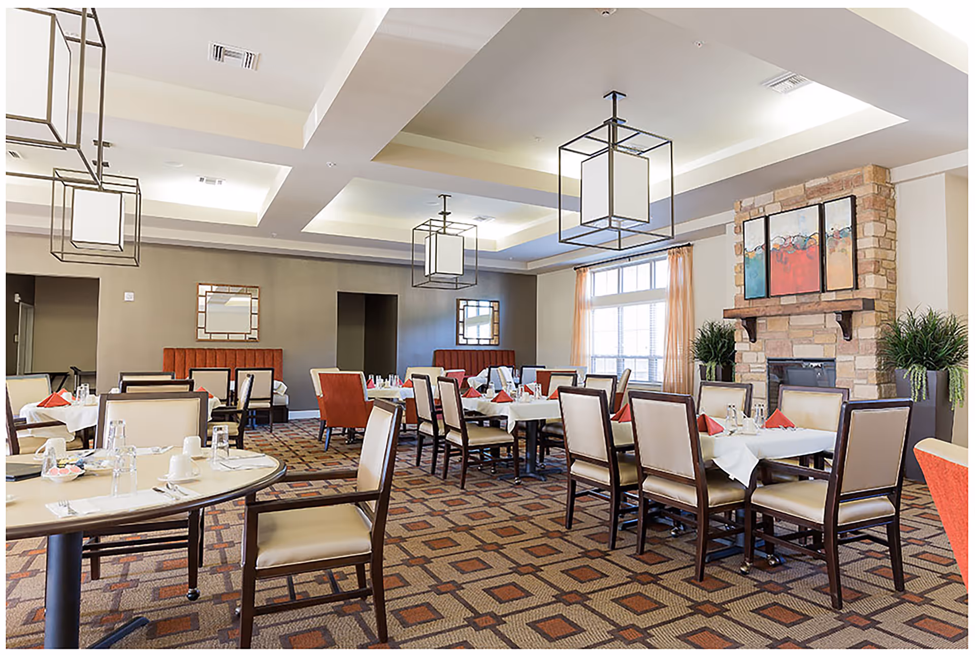 A spacious dining room with multiple tables set with white tablecloths, red napkins, and glassware. The room features beige walls, a patterned carpet, modern square pendant lights, and a stone fireplace with colorful artwork above it. There are large windows with light curtains and several chairs arranged around the tables.