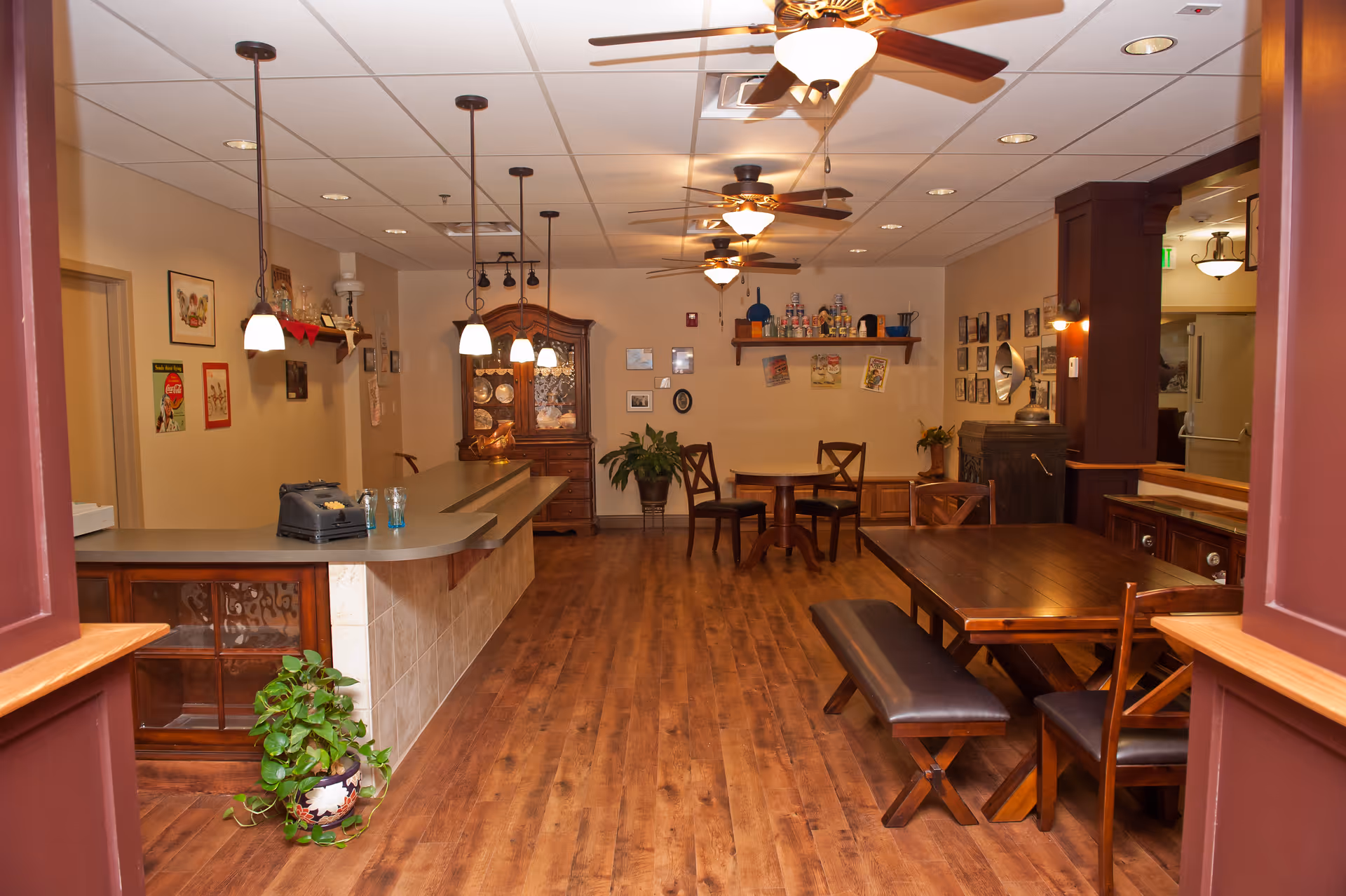 Interior view of a dining area in Colorow Care Center featuring wooden floors, a long counter with pendant lights, a wooden dining table with chairs and benches, ceiling fans with lights, and various decorative items on the walls and shelves.