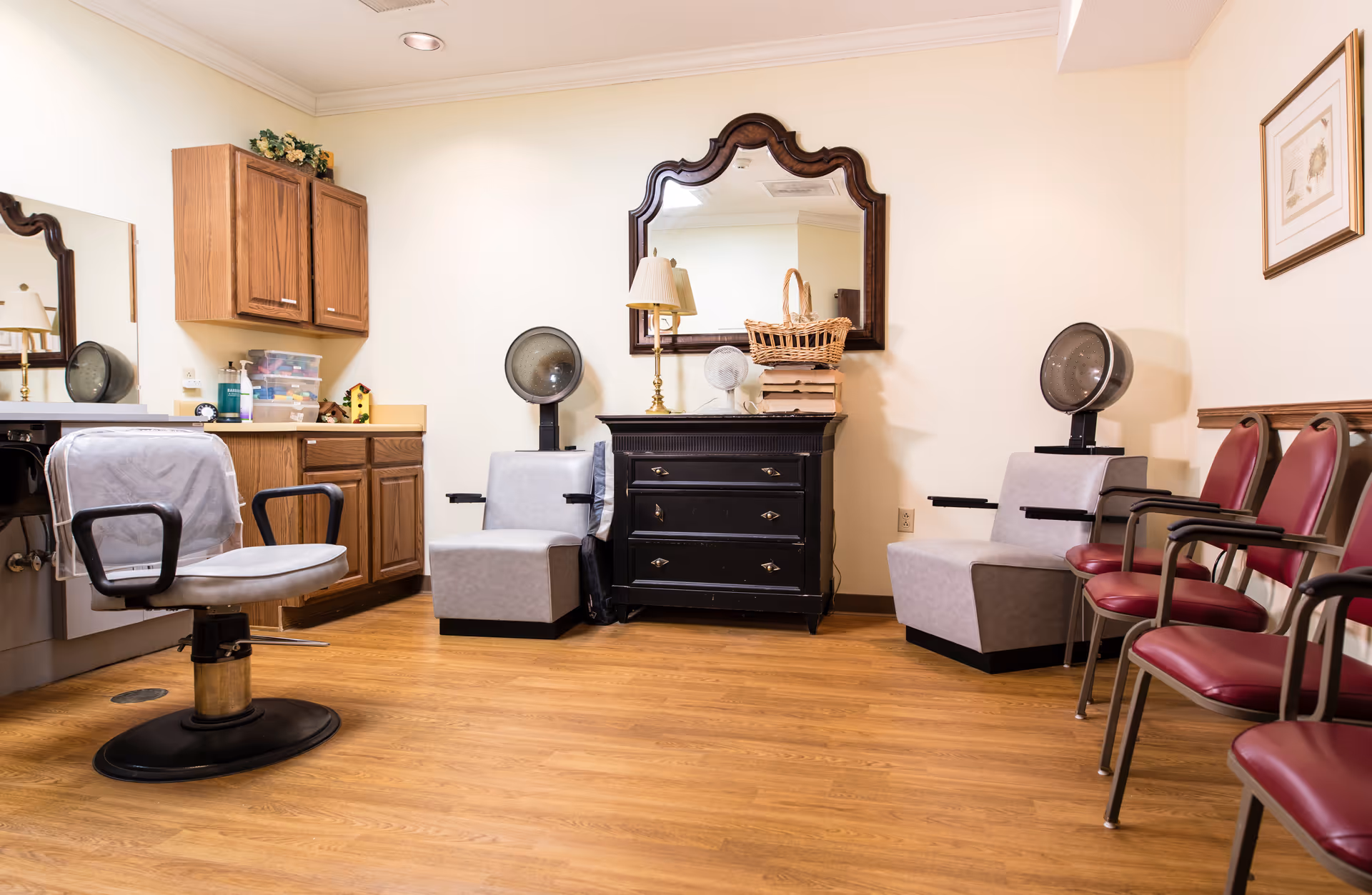 Interior salon area with styling chairs, hooded hair dryers, mirrors, cabinetry, and rows of waiting chairs on wood flooring.