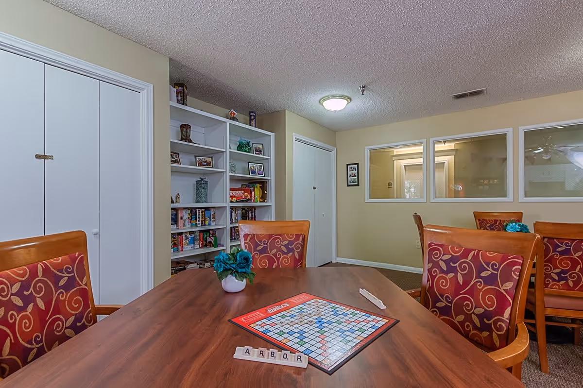 Community activity room with a wooden table and chairs, a Scrabble board on the table, bookshelves and interior windows.