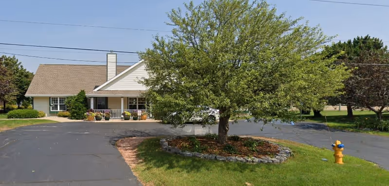 Front exterior view of a single-story building with a porch, surrounded by a paved driveway and landscaped greenery including a tree in the center and a yellow fire hydrant on the right side.
