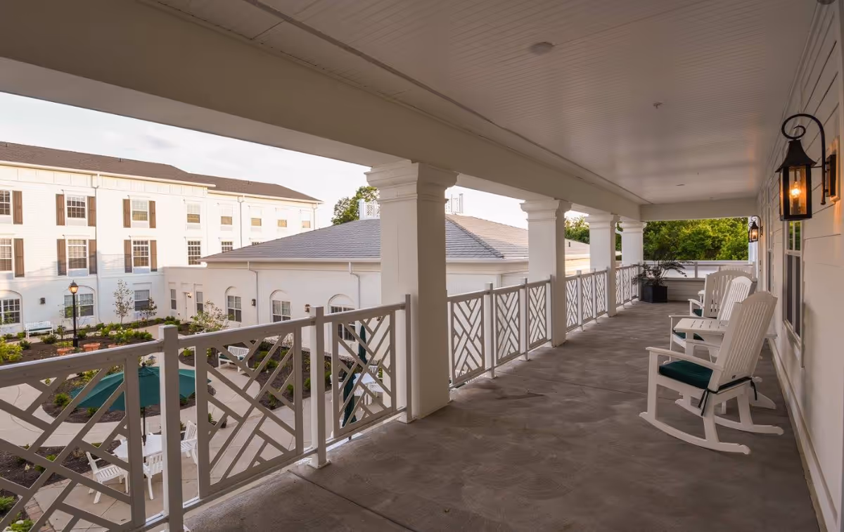 Covered outdoor balcony area with white rocking chairs featuring green cushions, white railing with geometric patterns, and hanging lantern-style lights. The balcony overlooks a courtyard with garden beds, benches, and a building with white walls and brown shutters.