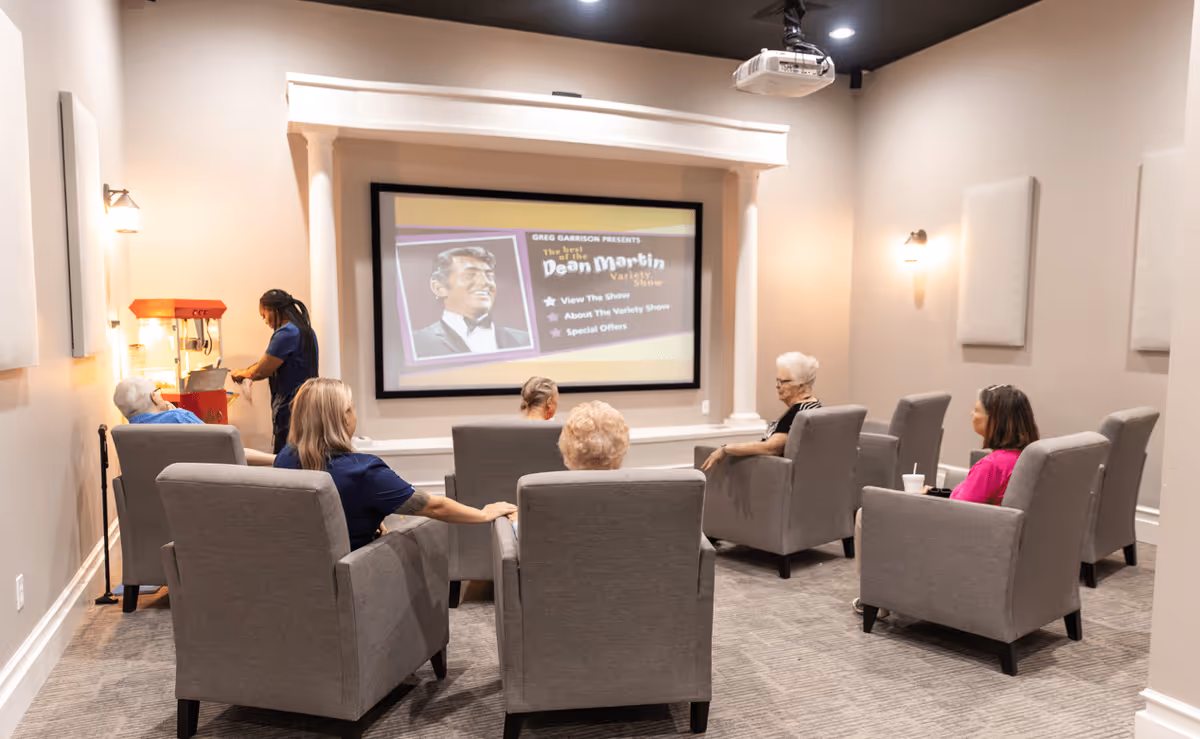 A group of elderly people seated in comfortable gray armchairs watching a movie or show projected on a large screen in a cozy room. A staff member is preparing popcorn at a popcorn machine on the left side of the room. The screen displays an image of Dean Martin with text about a variety show.