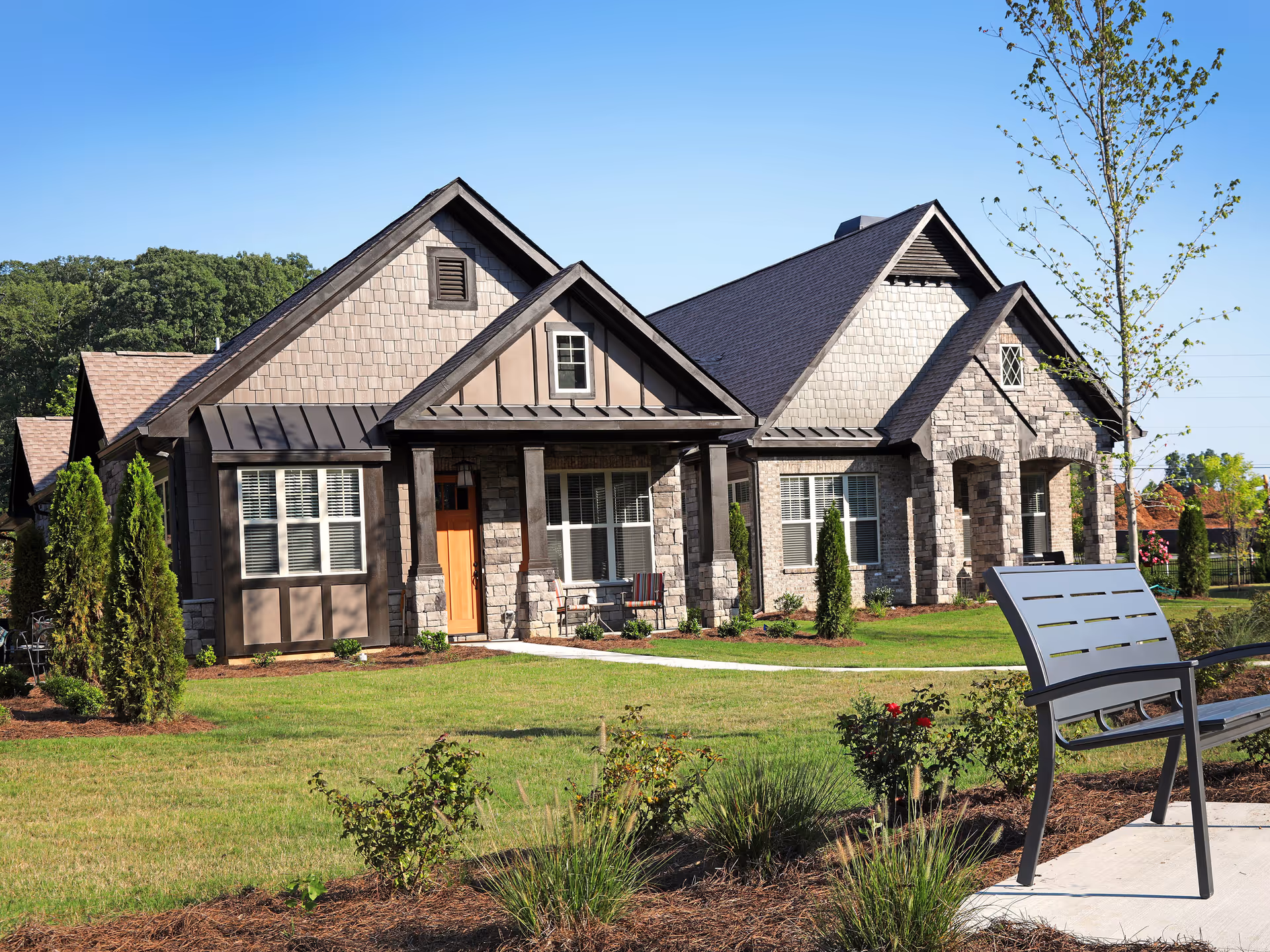 Exterior view of a single-story residential building with stone and wood siding, multiple gabled roofs, and a front porch. The building is surrounded by a well-maintained lawn, small trees, and shrubs. A metal bench is placed on a concrete path in the foreground under a clear blue sky.