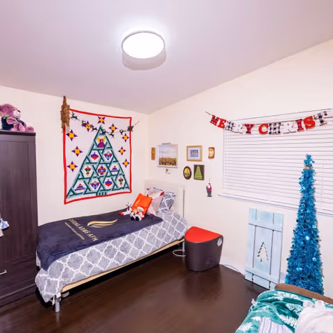 A decorated bedroom with a single bed covered in a gray and white patterned blanket and a dark blue blanket with the Caring Arms Adult Family Homes logo. The wall above the bed has a colorful quilt with a Christmas tree design and some hanging decorations. There is a small red and black stool next to the bed, a window with white blinds, and a blue Christmas tree decoration in the corner. Various small framed pictures and holiday decorations are on the walls.